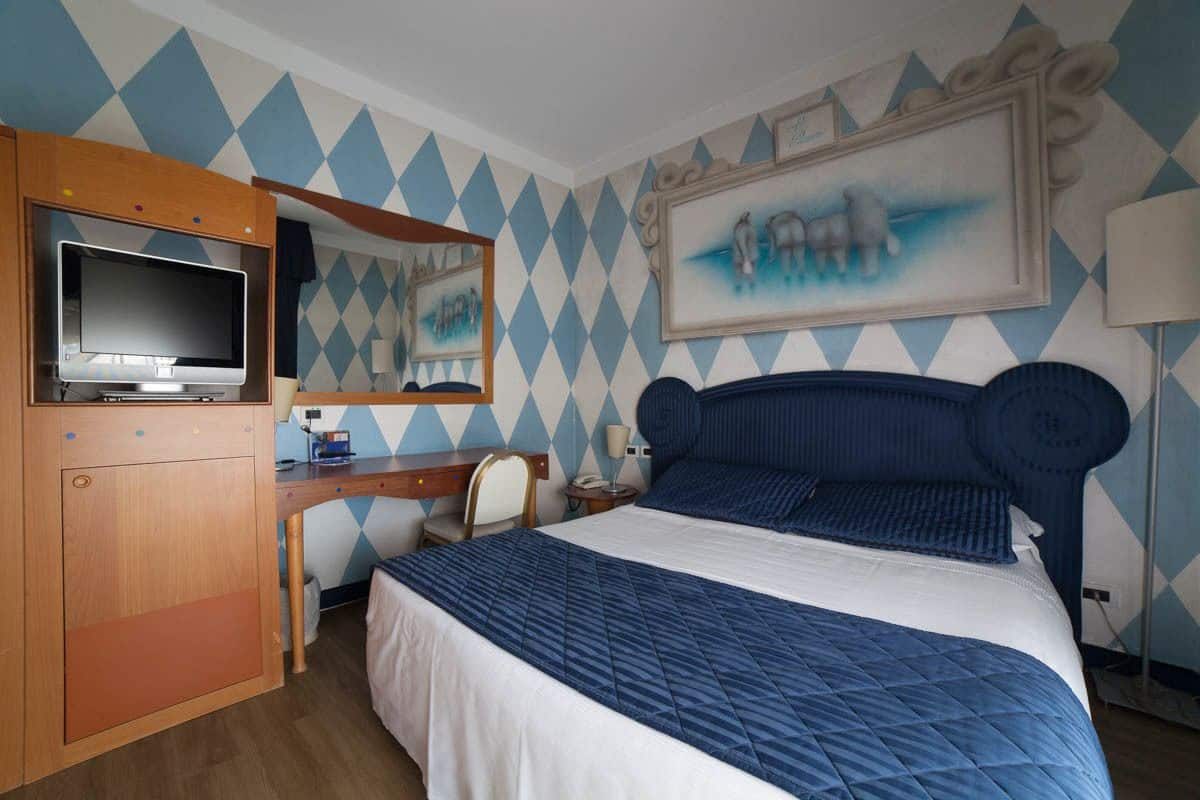 A hotel room with a bed, a desk, a TV, and a painting on the wall.