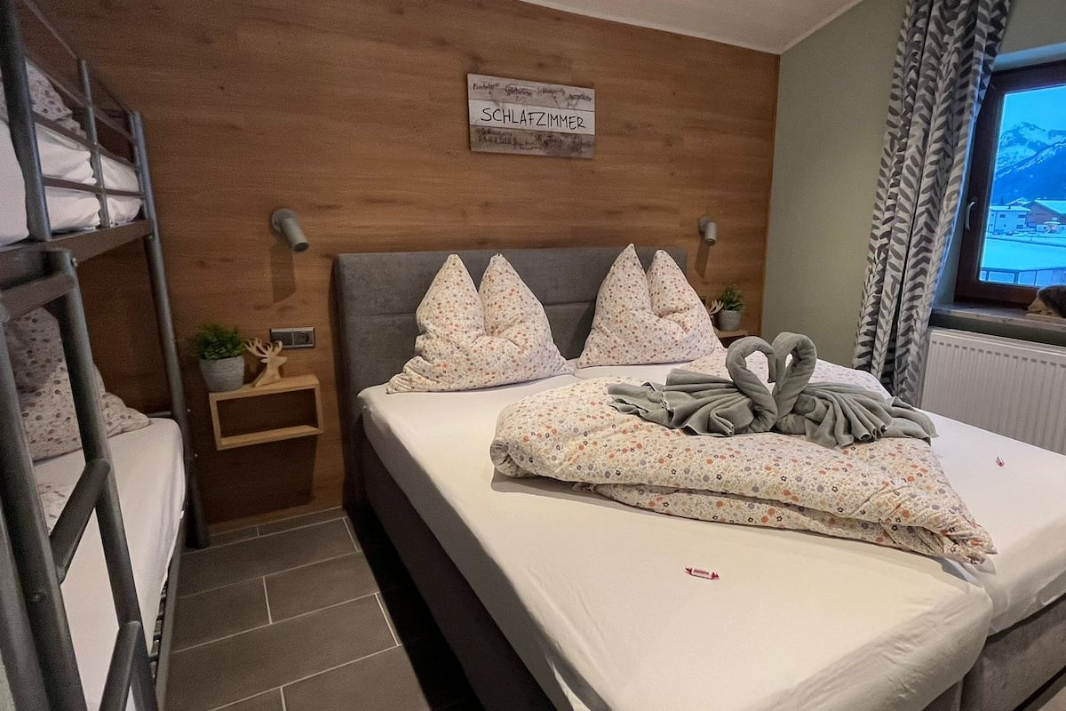 A bedroom with a bunk bed, a bed with floral bedding, a sign above the bed, a window with a view of mountains, and a plant on the nightstand.
