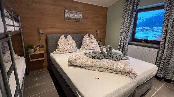 A bedroom with a bunk bed, a bed with floral bedding, a sign above the bed, a window with a view of mountains, and a plant on the nightstand. - Haus Tauern Am See (Heiterwang)