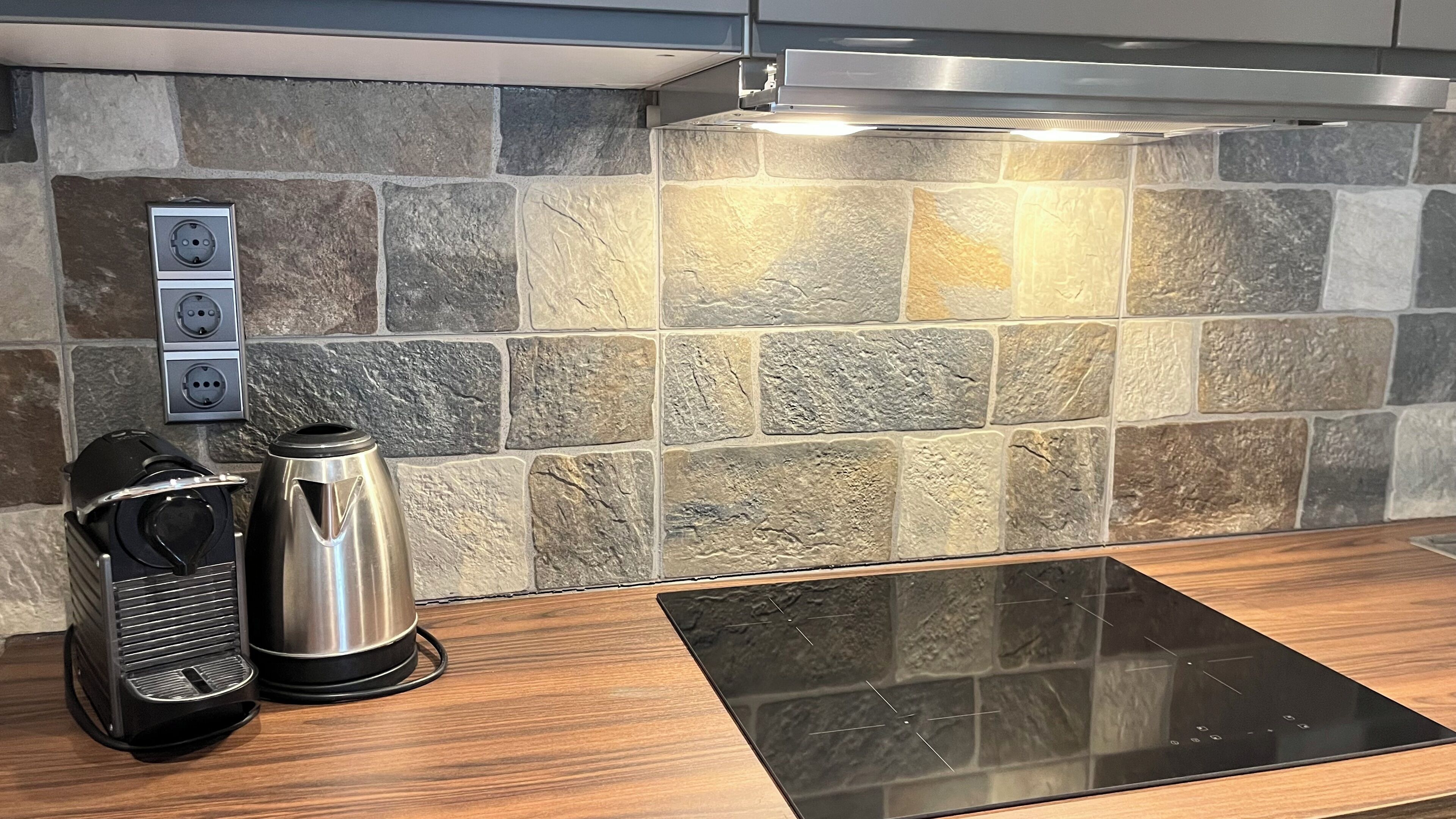 A kitchen with a stone backsplash, a coffee maker, and a kettle on a wooden countertop.