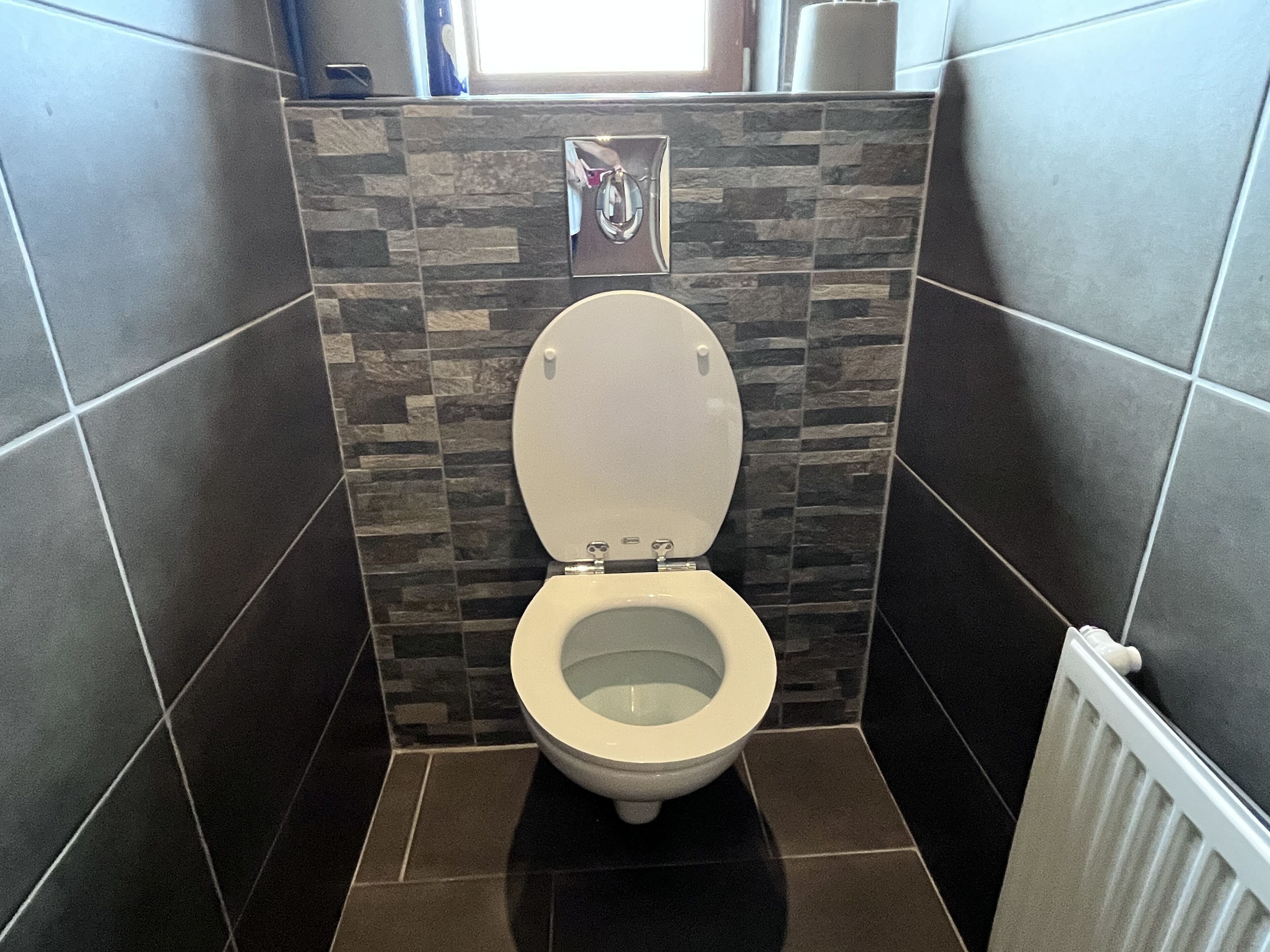 Ein Badezimmer mit Toilette, dunklen Fliesenwänden und einem Fenster über der Toilette.