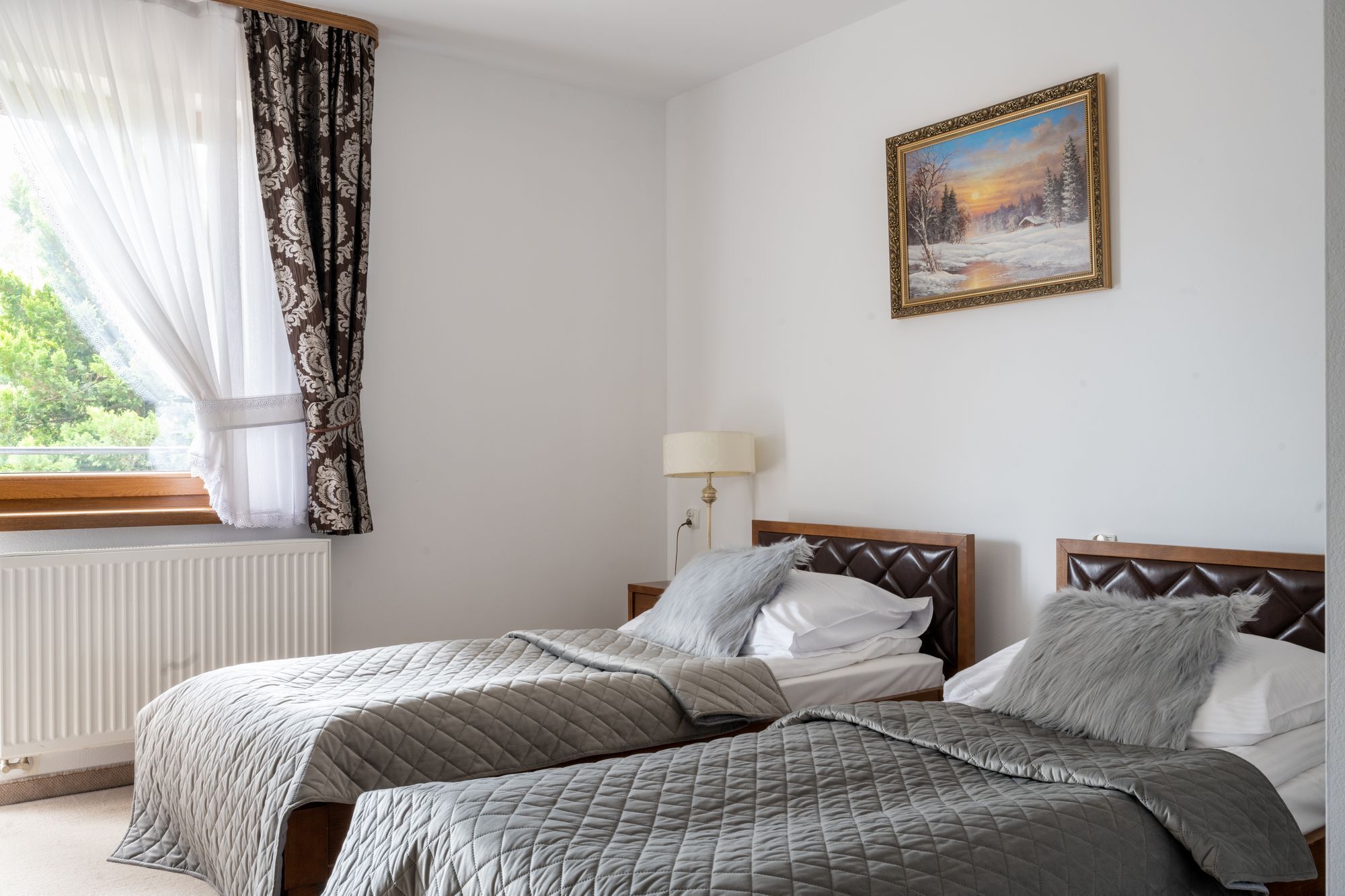 Un dormitorio con dos camas, una ventana con cortinas traslúcidas, un cuadro en la pared y una lámpara de noche.