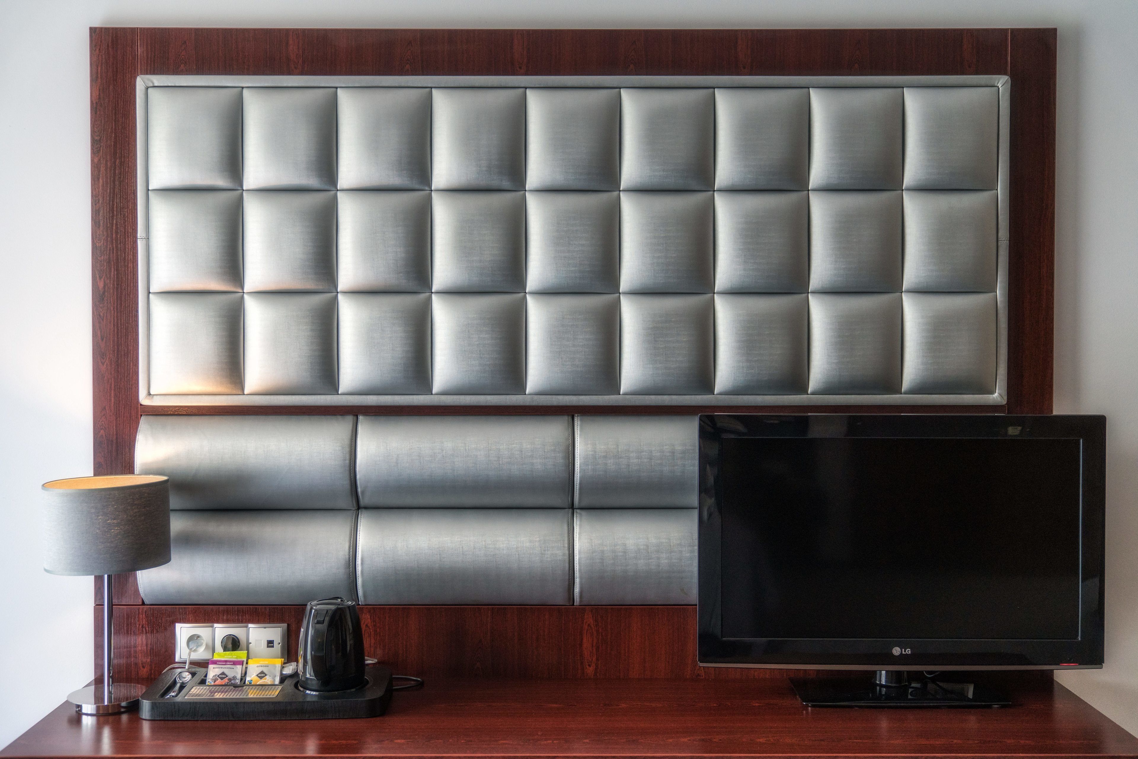 Habitación de hotel con un gran cabecero acolchado, un televisor de pantalla plana y una mesita de noche con lámpara y cafetera.