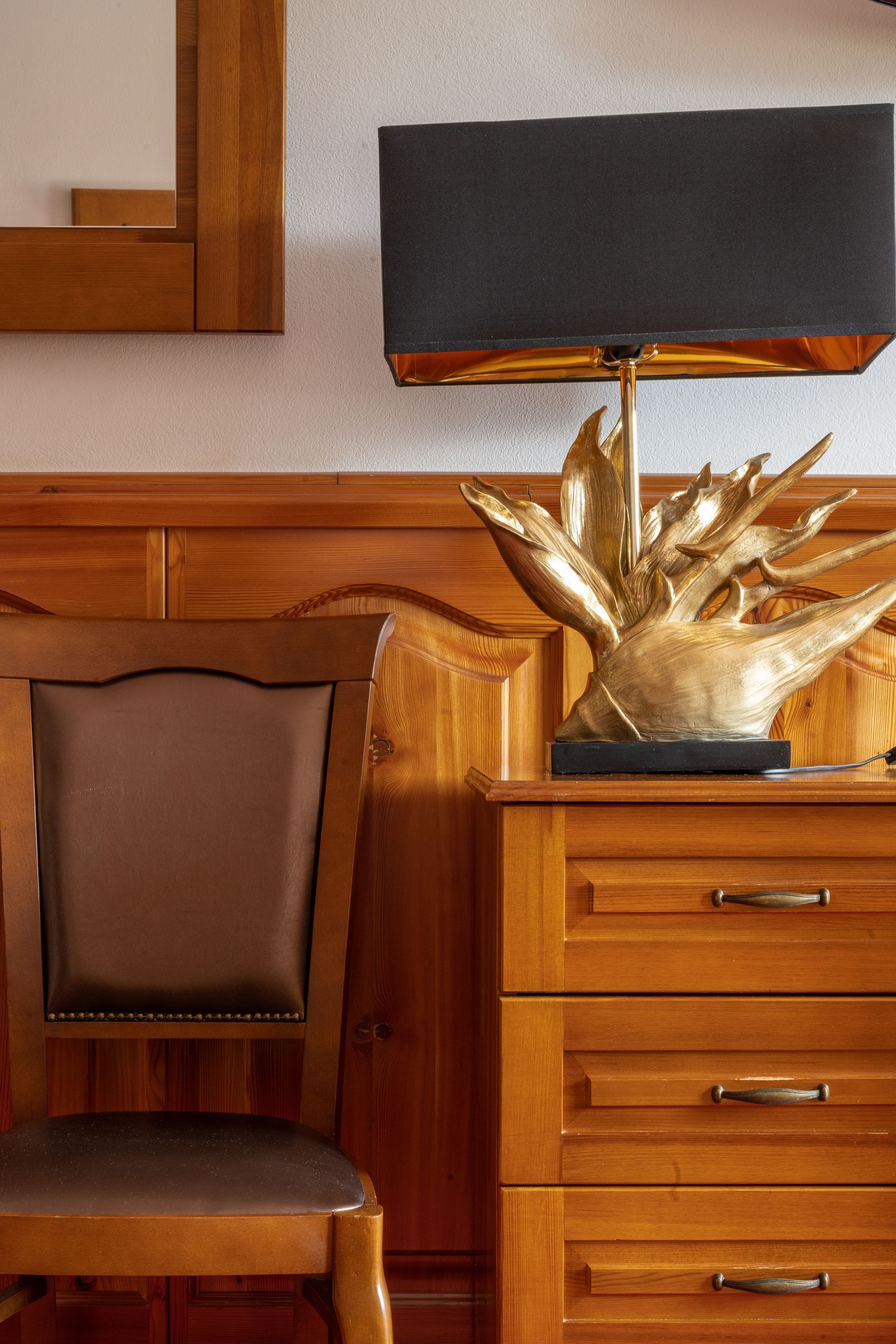 A wooden chair with a cushioned seat and backrest, a wooden dresser with drawers, and a table lamp with a black shade.