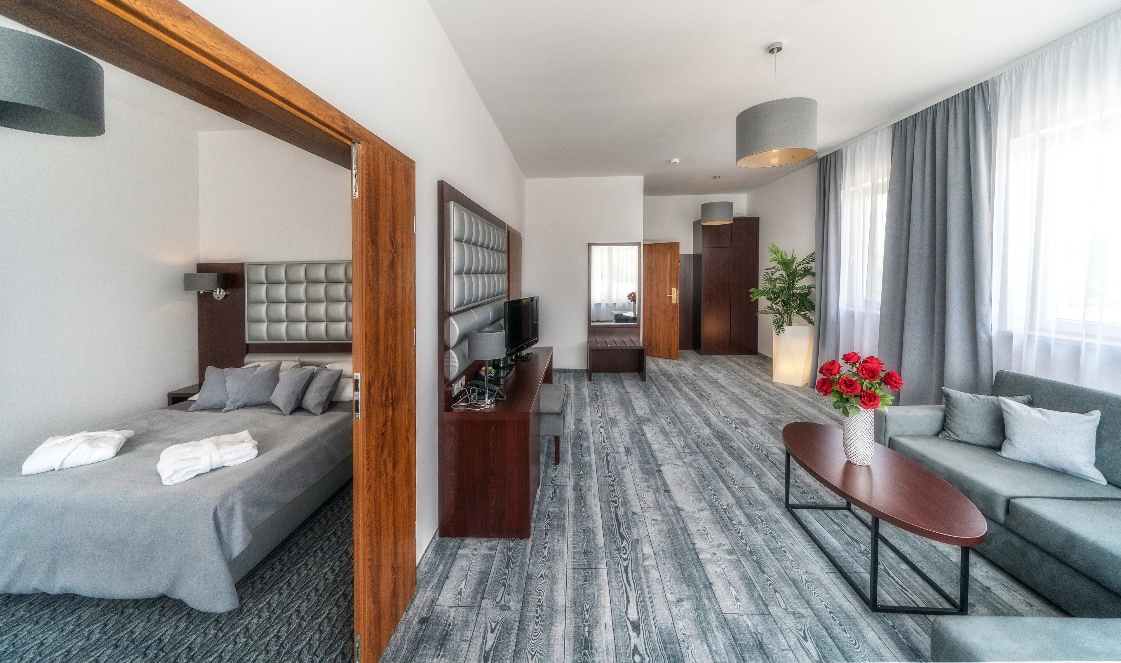 Quarto de hotel moderno com cama, área de estar com televisão, sofá e janela ampla com cortinas.