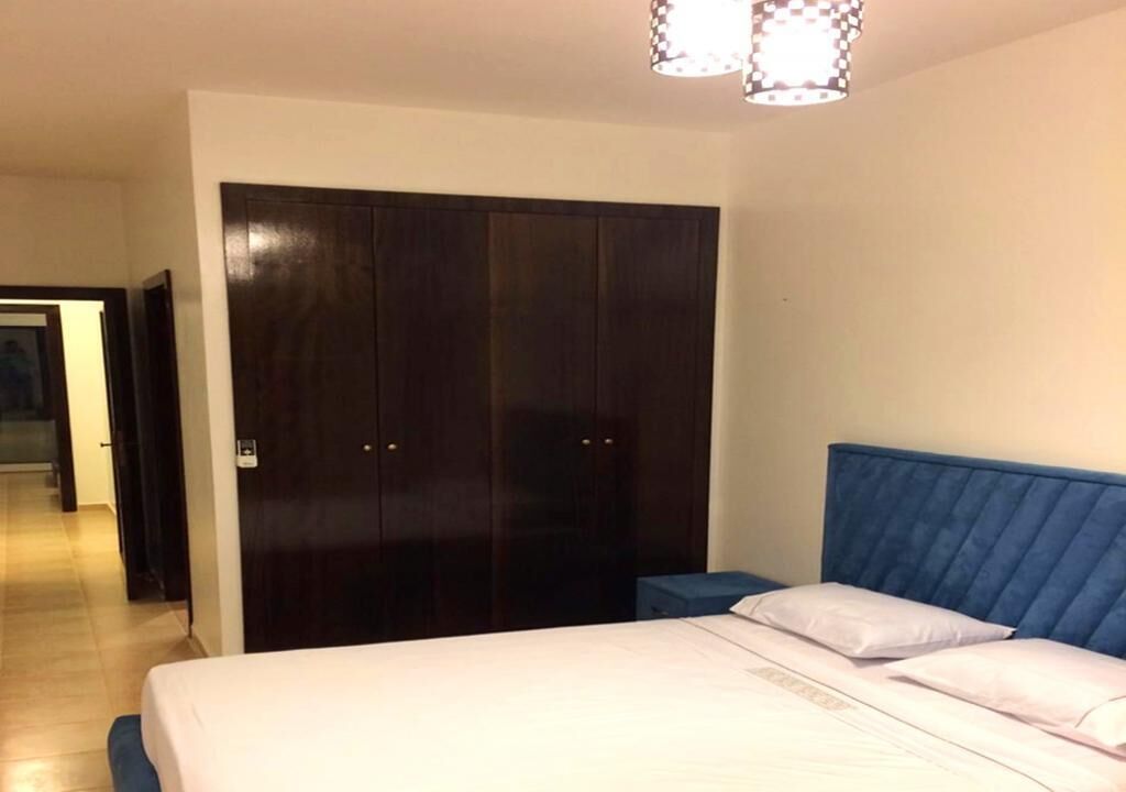 A bedroom with a large dark wooden wardrobe, a blue upholstered headboard, and a white bed with pillows.