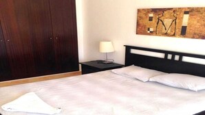 A neatly made bed with white linens, a wooden headboard, a nightstand with a lamp, and a painting on the wall. - Residence Marina Saidia 4239 (Saidia)