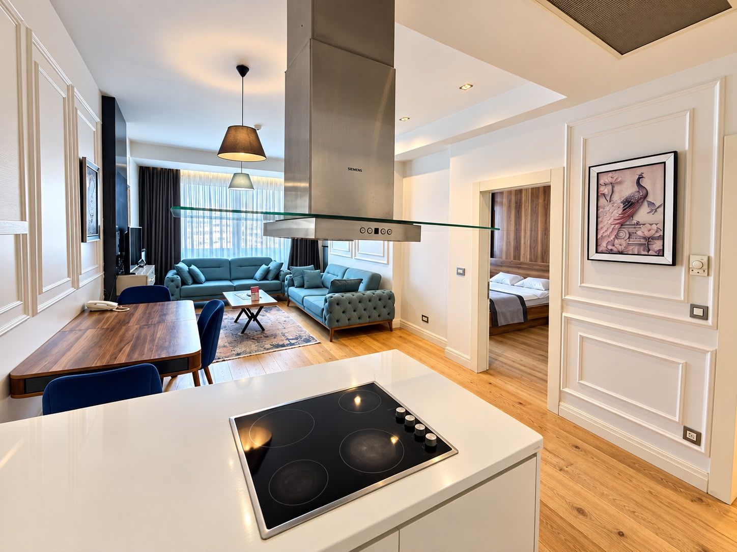 Uma cozinha moderna com fogão embutido, uma sala de jantar com mesa e cadeiras de madeira, uma sala de estar com sofá e mesa de centro, e um quarto com cama e uma moldura com imagem na parede.