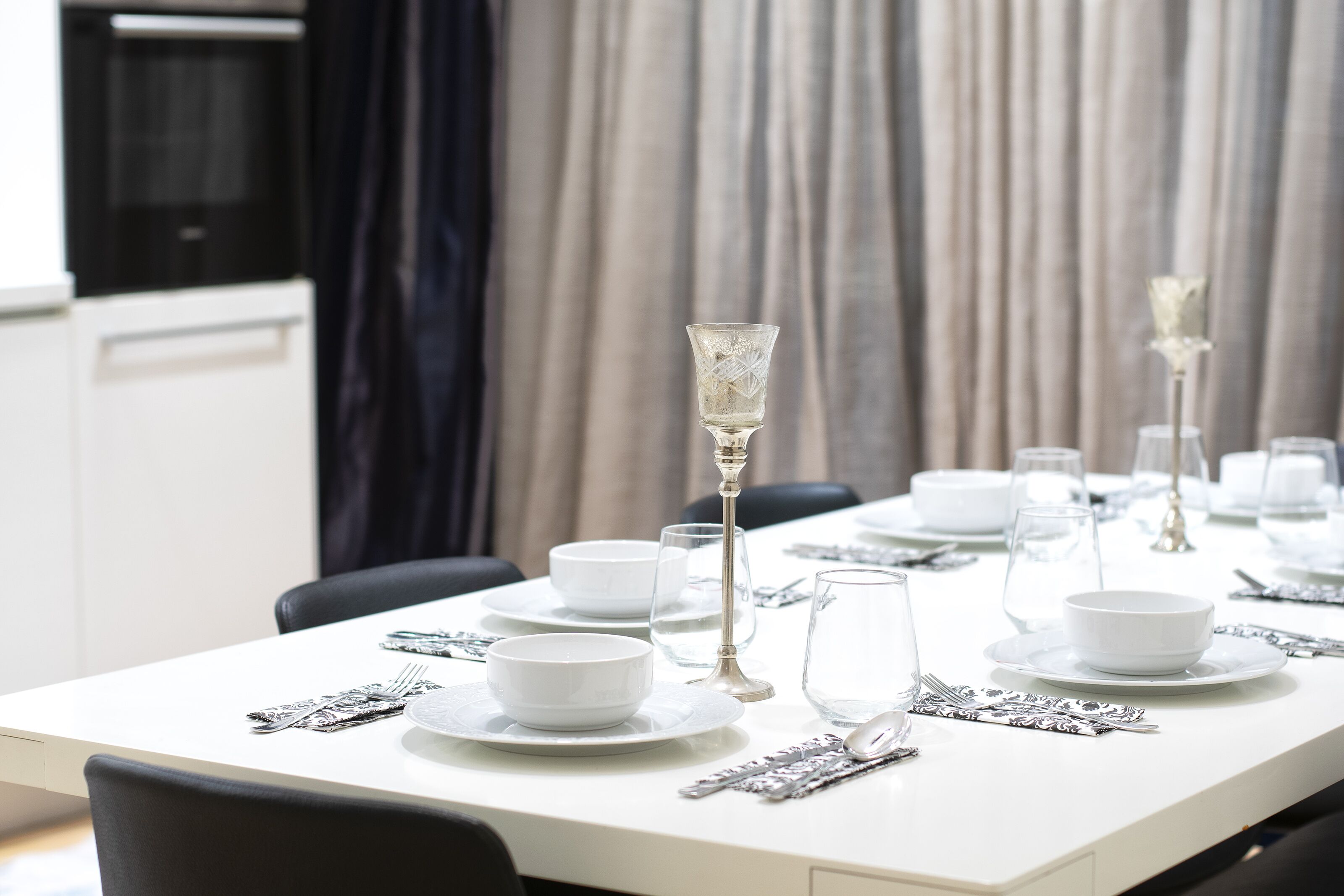 A neatly set dining table with white plates, bowls, and silverware, and a tall glass candle holder.