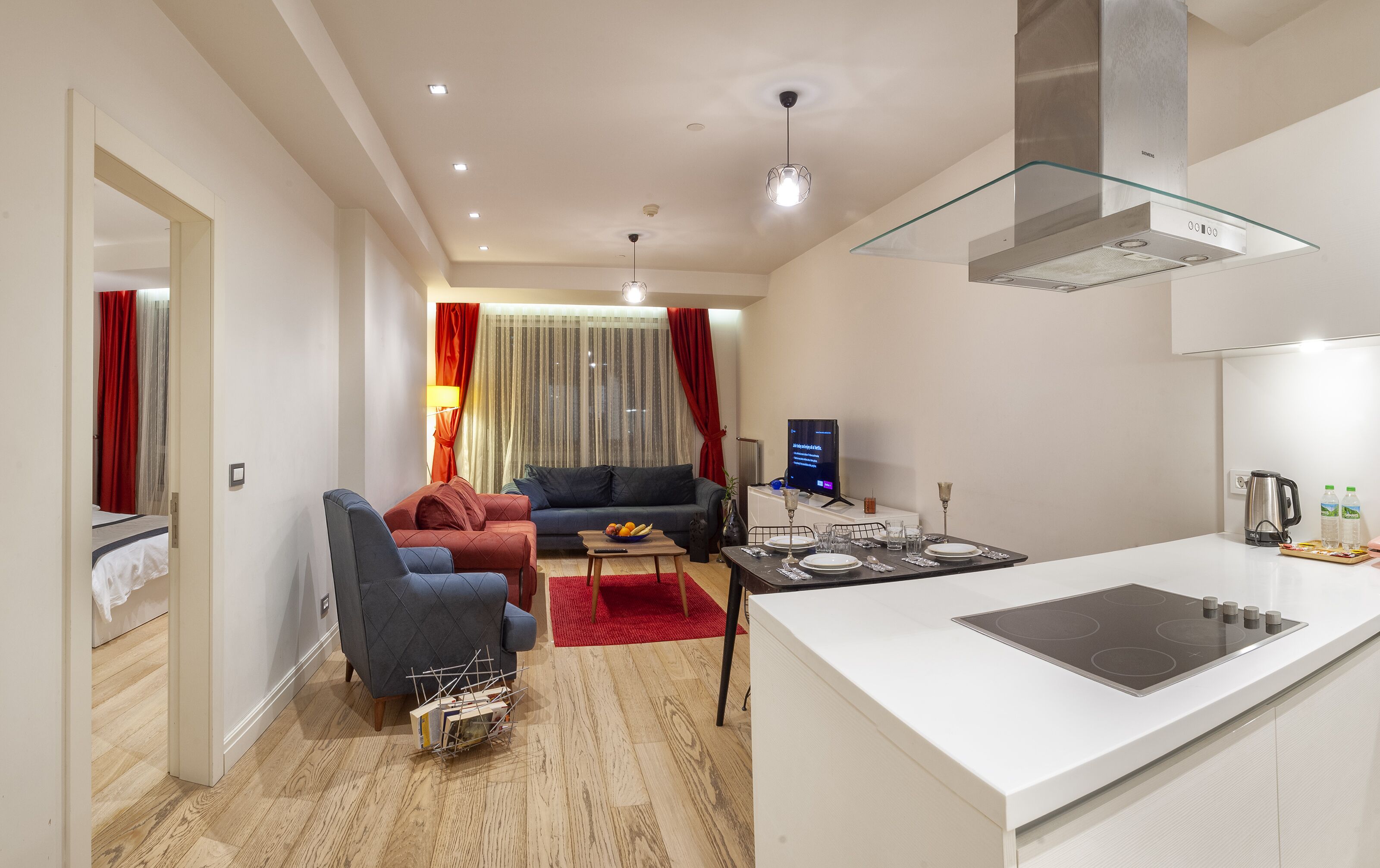 A modern living room with a sofa, a coffee table, and a TV. There is a kitchen area with a stove and a kettle.