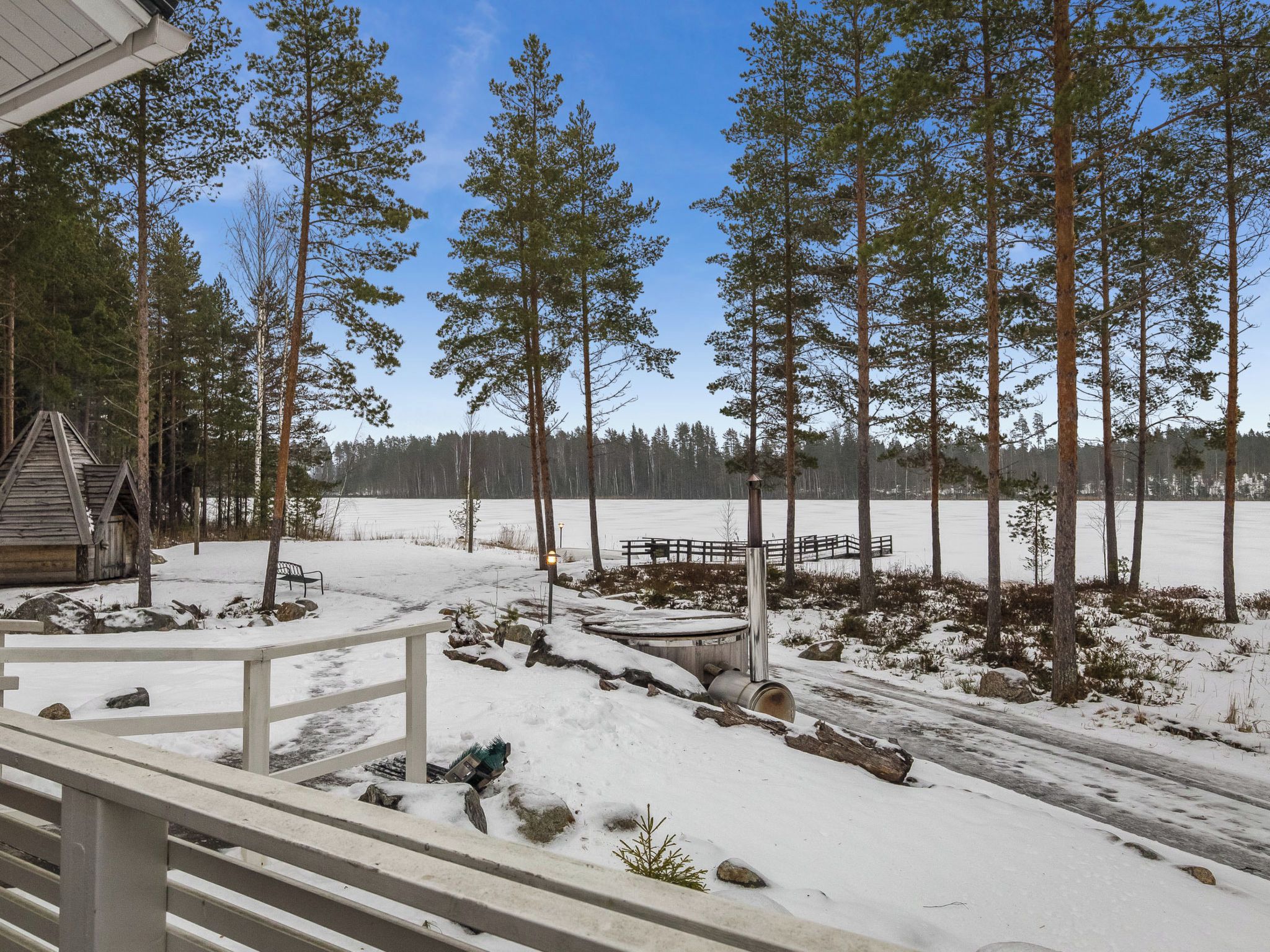 Lumipeitteinen maisema, jossa on puinen mökki, puinen terassi ja kirkas taivas.