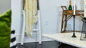 Una habitación con pared blanca, una escalera de madera apoyada en ella, una mesa con velas y una silla con cojín verde.