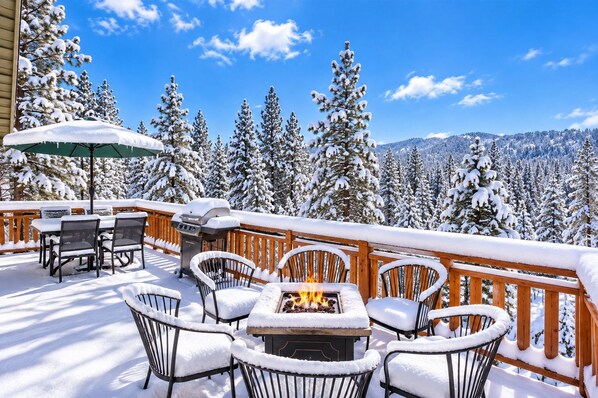 A snow-covered outdoor deck with a fire pit, chairs, and a table, surrounded by snow-laden trees and a clear sky. - View Point - Hot Tub + Slope Views | Game Room (Big Bear Lake)