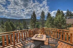 Une terrasse en bois avec un foyer et des sièges d’extérieur, entourée d’arbres et offrant une vue sur un paysage montagneux.