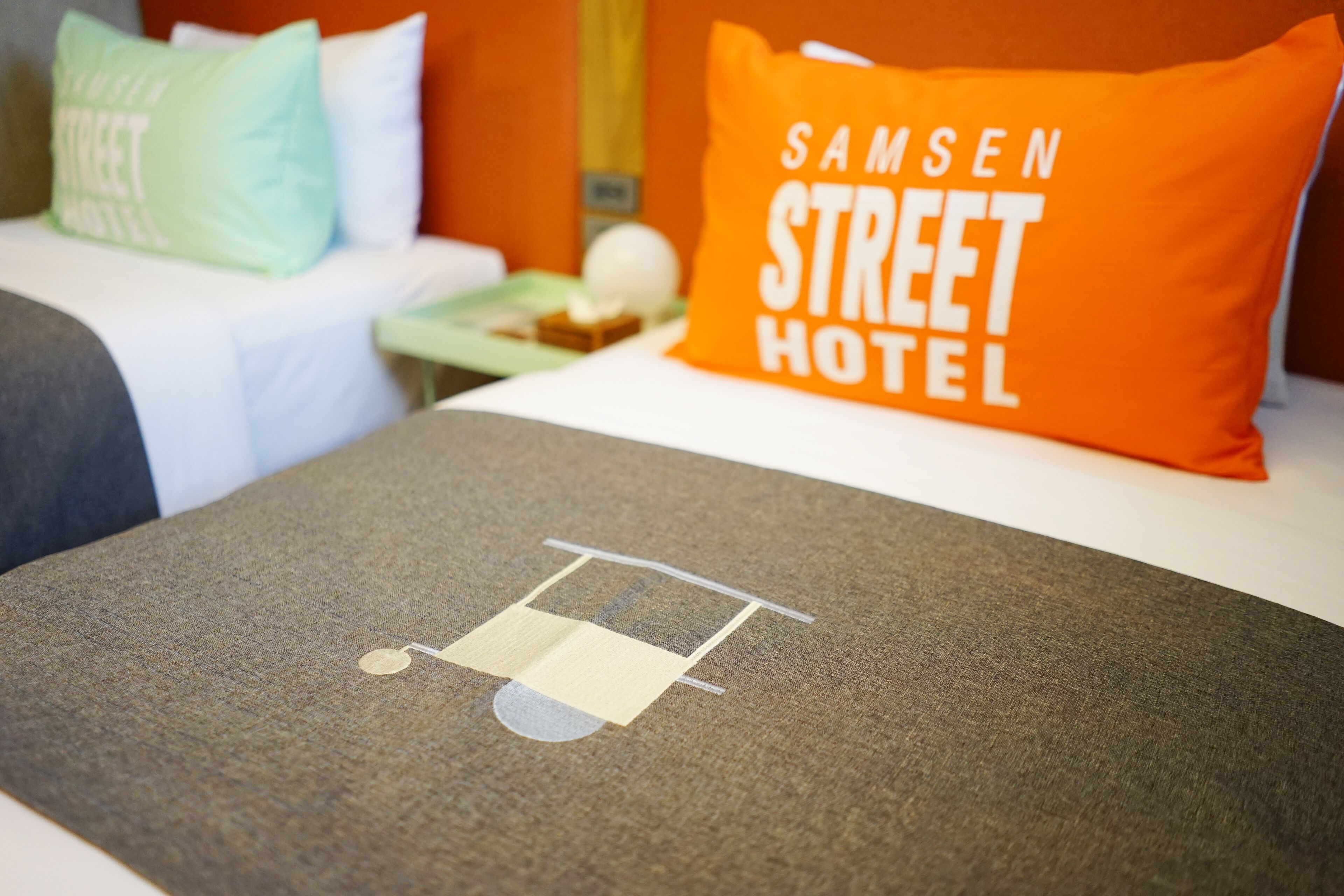A hotel room with a bed, a pillow with "SAMSEN STREET HOTEL" text, and a table with a small item on it.