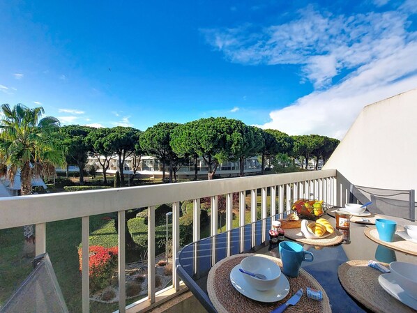 A balcony with a table set for breakfast, overlooking a garden with trees and a view of buildings. - La Frégate by Interhome (Port Camargue)