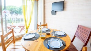 Zona pranzo con tavolo in legno apparecchiato per quattro persone, TV a parete e vista sull'esterno attraverso ampie finestre.