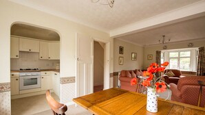 A dining area with a wooden table and chairs, a vase with flowers, and a kitchen in the background.