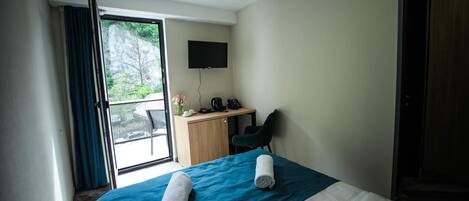 A hotel room with a bed, a desk, a chair, a TV, and a balcony with curtains.