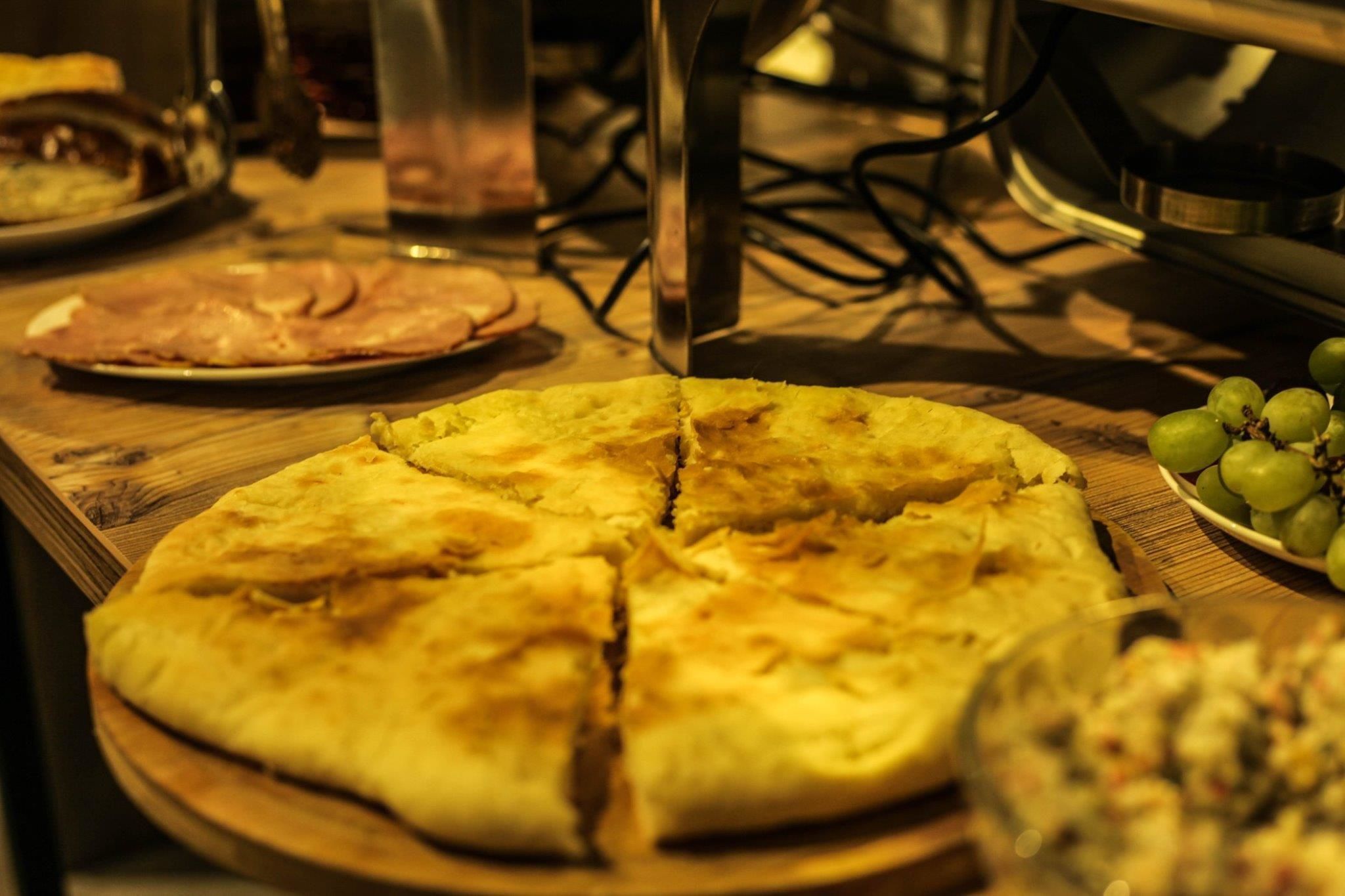 A pizza on a wooden board with a glass of water and a plate of sliced ham in the background.