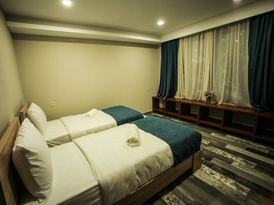 Una habitación de hotel con dos camas, un banco de madera, una ventana con cortinas y una alfombra estampada.