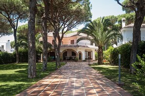 Un chemin menant à un bâtiment doté d’arches au niveau des fenêtres et d’un toit recouvert de tuiles, entouré d’arbres et de verdure.