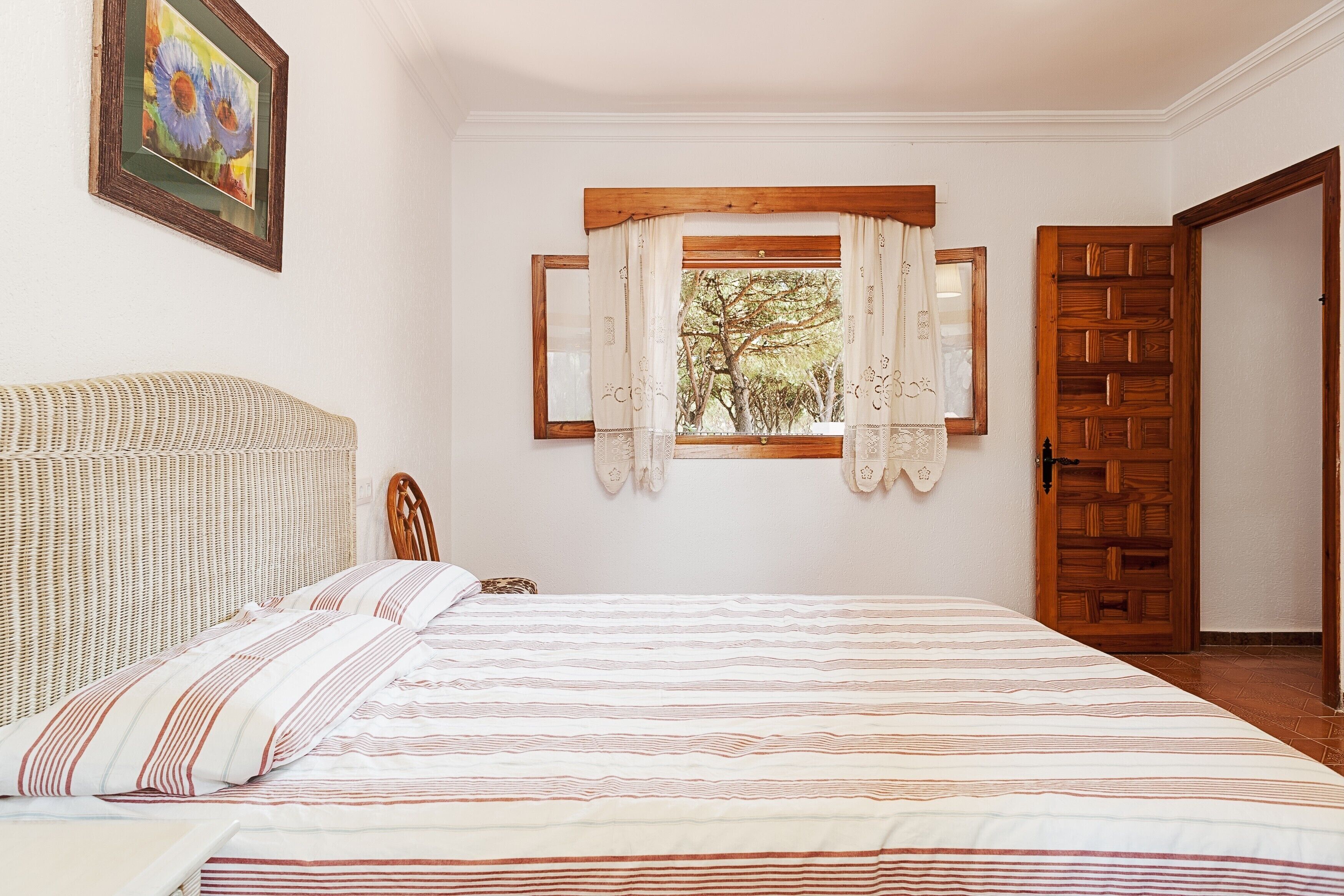 Un dormitorio con cama, ventana con cortinas, puerta de madera y un cuadro enmarcado en la pared.
