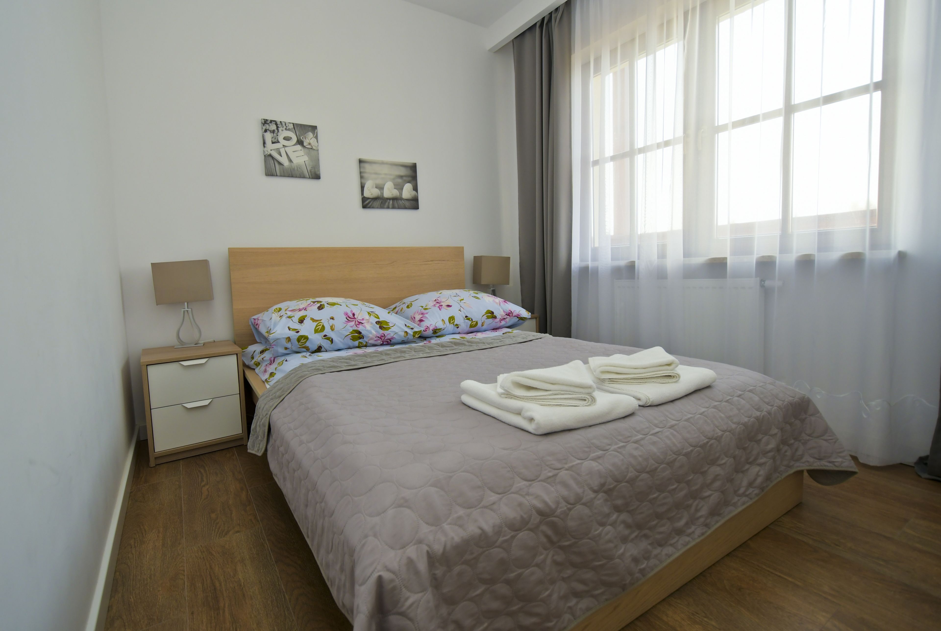 Ein ordentlich bezogenes Bett mit einer gesteppten Decke, ein Nachttisch mit Lampe und ein Fenster mit transparenten Vorhängen.