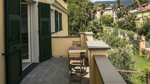 Un balcon avec une table et des chaises, une porte avec des volets et une vue sur un quartier résidentiel, avec des arbres et des maisons.