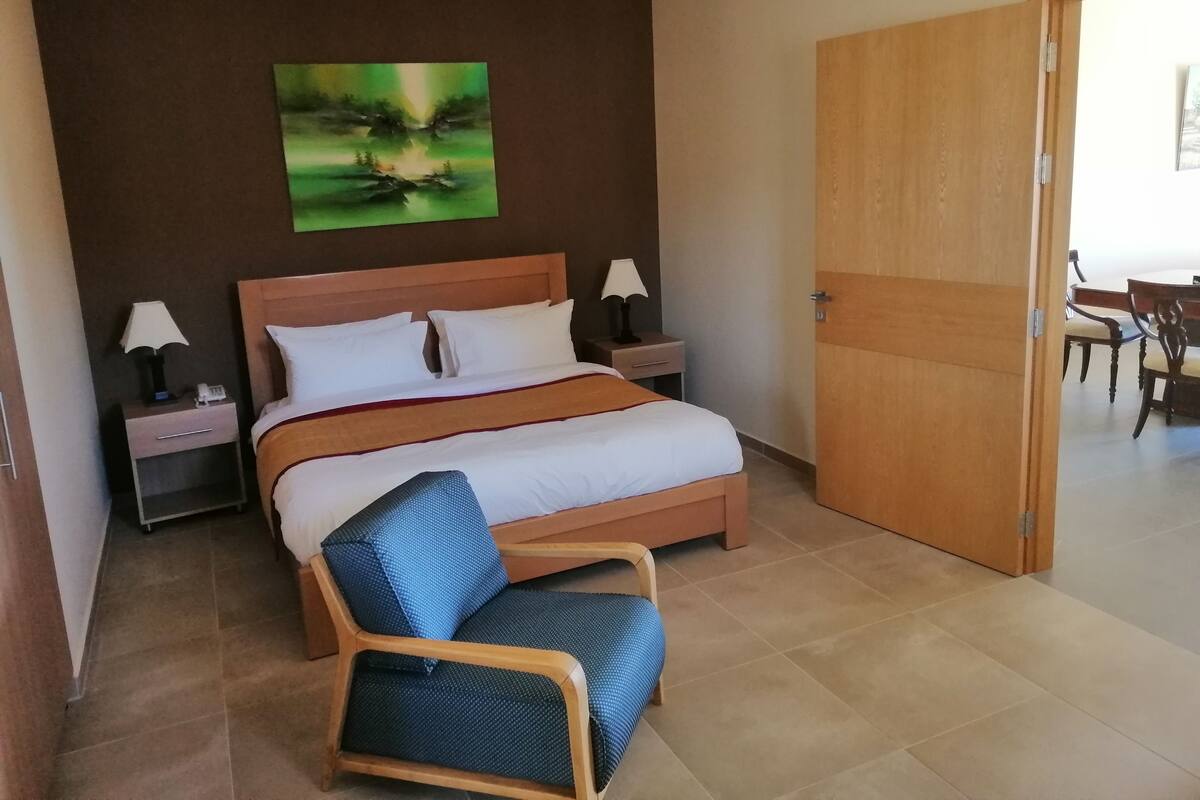 A hotel room with a bed, two bedside tables with lamps, a wooden chair, and a dining area visible through an open door.