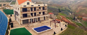 Un edificio de varios pisos con piscina y zona de estar al aire libre, rodeado de un paisaje pintoresco.