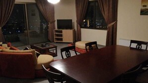 A living room with a sofa, ottoman, and a dining table with chairs. There is a TV on a stand and a window with curtains. - Zahle Hills Hotel (Zahlé)