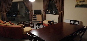 Una sala de estar con sofá, reposapiés y una mesa de comedor con sillas. Hay un televisor sobre un mueble bajo y una ventana con cortinas.