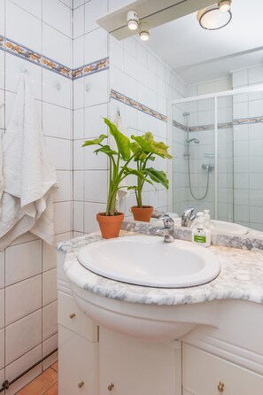 Un baño con lavamanos blanco, un espejo, ducha y una planta en maceta.