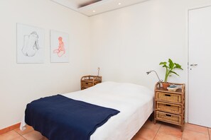 A bedroom with a bed, two wall-mounted artworks, a wicker side table with a plant, and a white door.