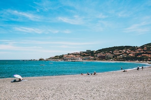 A pebble beach with a clear blue sea, buildings on a hillside, and people relaxing.