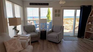 A living room with two armchairs, a side table, a lamp, a window with a view of the beach, and a sign reading 'Another Day In Paradise'. - Stay right on the beach.  Enjoy the view from our updated home right on the Gulf (Gulf Shores)