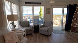 A living room with two armchairs, a side table, a lamp, a window with a view of the beach, and a sign reading 'Another Day In Paradise'.