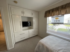 A bedroom with a TV on a white cabinet, a window with blinds, and a bed.