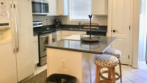 A kitchen with white cabinets, a refrigerator, a microwave, a stove, and a sink. There is a clock on the wall and a window with blinds.