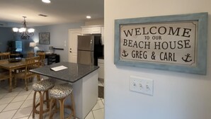 A kitchen with a central island, wooden chairs, and a sign that reads "WELCOME TO OUR BEACH HOUSE GREG & CARL".