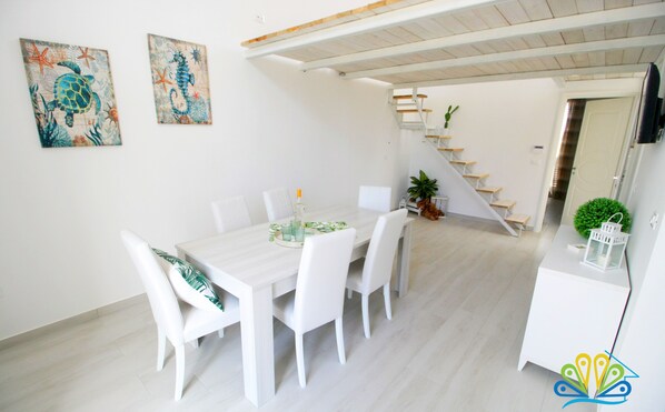 Un'area pranzo con un tavolo e sedie bianche, una scala e una piccola pianta su un mobile bianco.