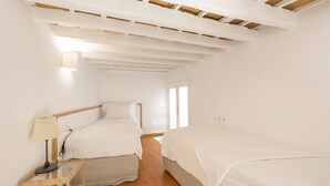 Un dormitorio con dos camas, suelo de madera, una mesita de noche con lámpara y una ventana que deja entrar luz natural.