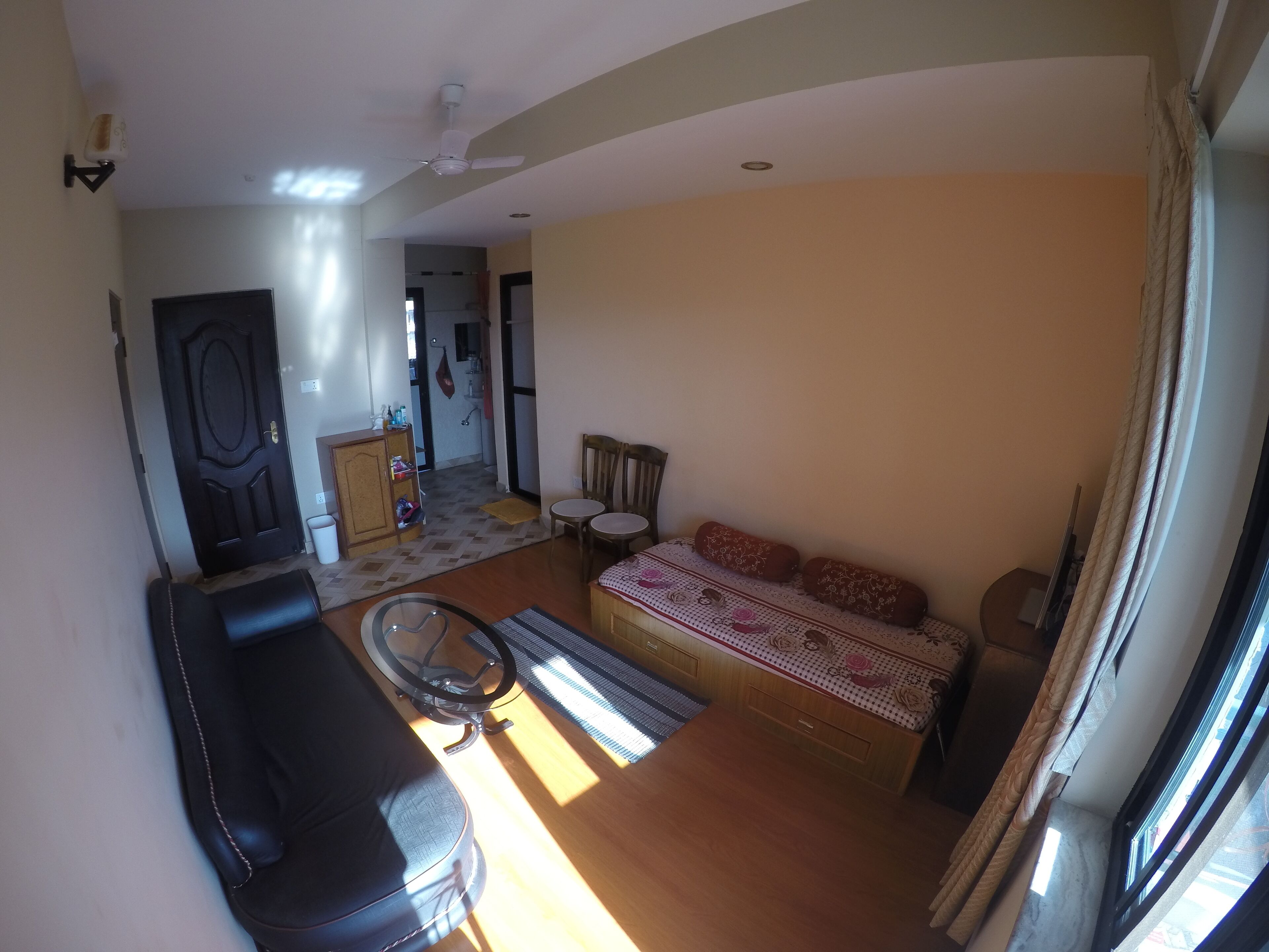 En stue med sofa, et lille bord, en seng med mønstret sengetøj, en loftventilator og et vindue med gardiner.