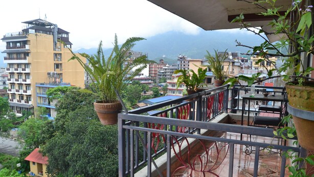 A balcony with potted plants, a tiled floor, and a view of surrounding buildings and mountains. - Ranss Apartment (Pokhara)