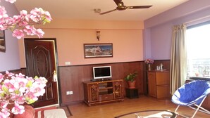 A living room with a TV, wooden furniture, a ceiling fan, a potted plant, and a balcony with a view. - Ranss Apartment (Pokhara)