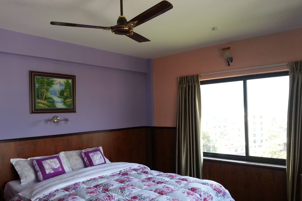 A hotel room with a bed, a ceiling fan, a window with curtains, and a painting on the wall.