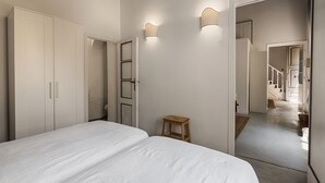 Un dormitorio con una cama grande, un armario, un taburete de madera y un espejo que refleja una escalera.
