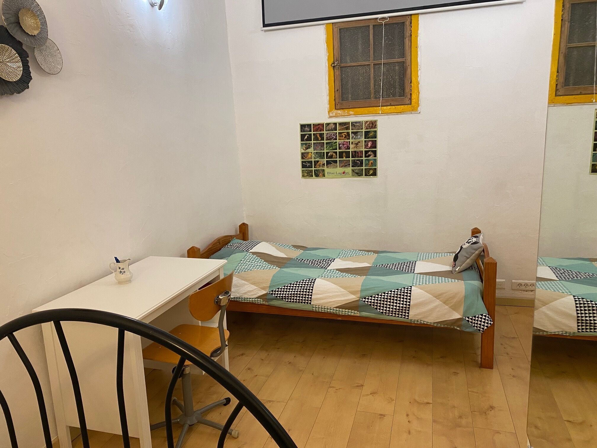 Een kleine, knusse kamer met een enkel bed, een bureau en een stoel. Op de muur is een klein plankje met decoratieve spullen te zien.