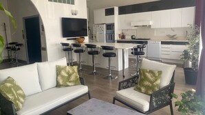 A modern living room with a sofa, chairs, and a coffee table. A kitchen is visible in the background with a TV mounted on the wall. - Large apartment on canal 2 steps from downtown Sète (Sète)
